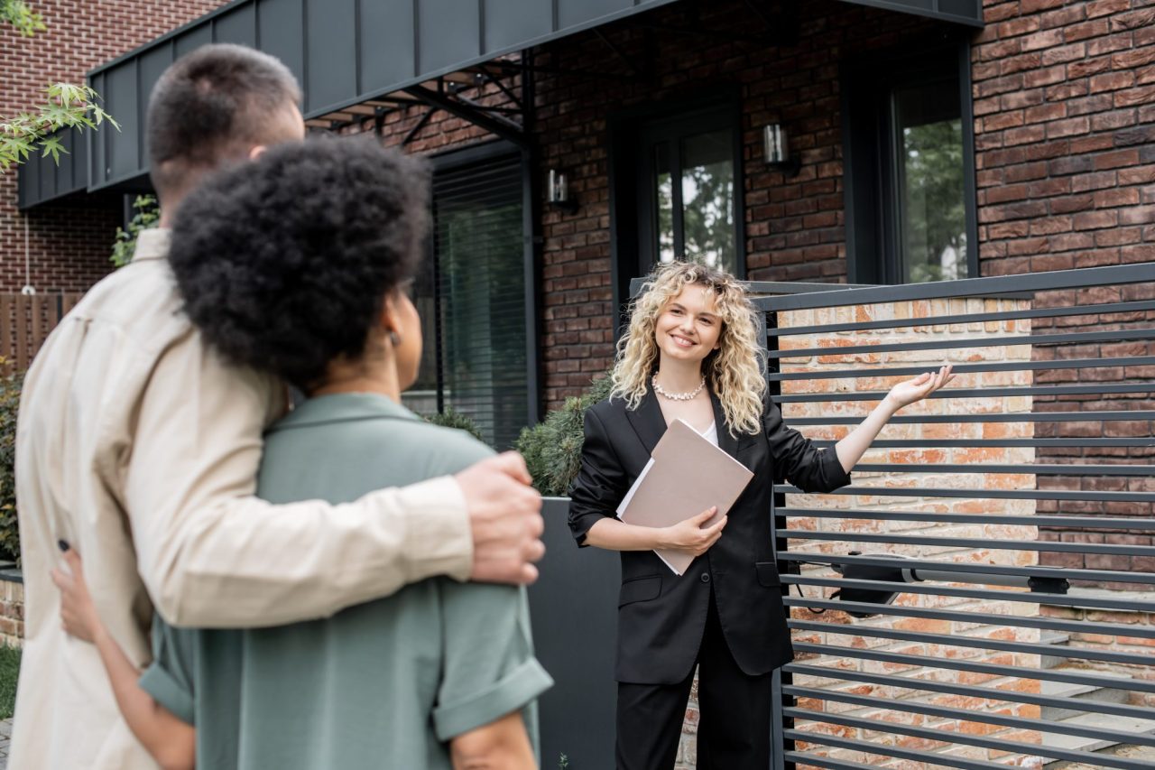 cheerful property agent with folder showing new city cottage to multiethnic couple of buyers